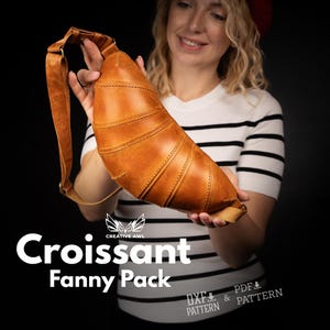 May include: A brown leather fanny pack shaped like a croissant, with a long strap. The bag has stitched details and is held by a person. The text "Croissant Fanny Pack" is visible in white.