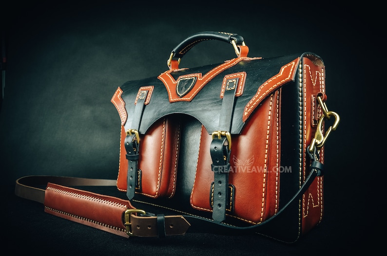 May include: A handcrafted leather briefcase in black and brown with white stitching. The bag features two front pockets, a top handle, and a detachable shoulder strap. Gold-colored hardware accents the design.