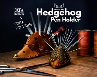 May include: A leather hedgehog pen holder with a brown body and green accents. The holder is filled with sewing needles and a small awl. The image also includes spools of thread in various colors and a pair of scissors.
