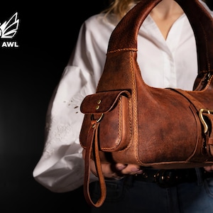Puede incluir: Un bolso de mano de cuero marr&oacute;n de estilo satchel con cierre de solapa y hebilla. El bolso tiene un bolsillo frontal y una correa larga.