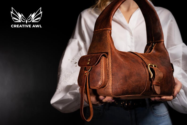 Puede incluir: Un bolso de mano de cuero marr&oacute;n de estilo satchel con cierre de solapa y hebilla. El bolso tiene un bolsillo frontal y una correa larga.