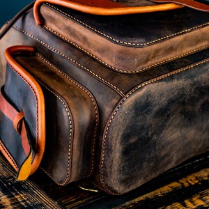 May include: A brown leather backpack with a flap closure and a front pocket. The backpack has orange stitching and a distressed look.
