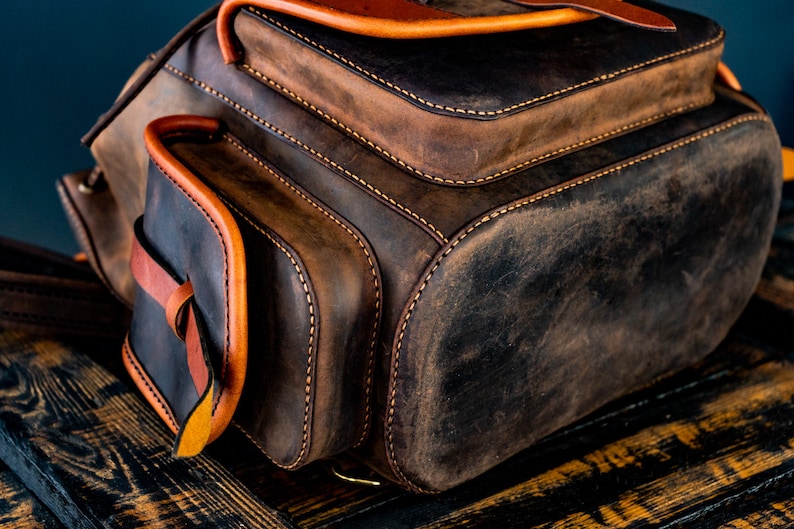 May include: A brown leather backpack with a flap closure and a front pocket. The backpack has orange stitching and a distressed look.