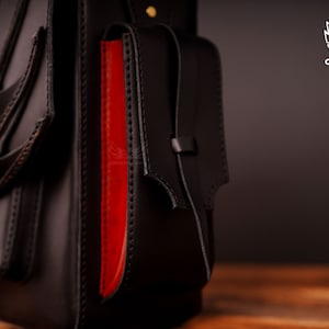 Op de afbeelding: Een zwarte leren tas met rode accenten en een klep sluiting. De tas heeft een decoratief vleugelontwerp aan de voorkant en een klein vakje aan de zijkant.