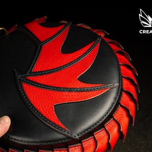 May include: A round handbag in black and red with a distinctive pattern. The bag has a black body with a red, stylized design on the front. The edges are trimmed with a red, textured material. The Creative Awl logo is visible.