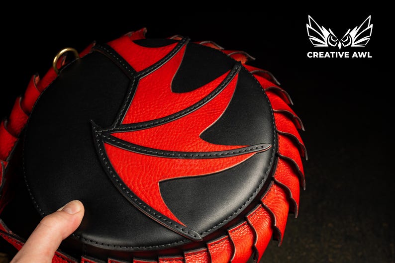 May include: A round handbag in black and red with a distinctive pattern. The bag has a black body with a red, stylized design on the front. The edges are trimmed with a red, textured material. The Creative Awl logo is visible.