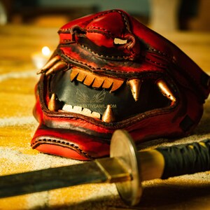 K&ouml;nnte beinhalten: Eine rote Leder-Samurai-Maske mit goldenen Akzenten und scharfen Z&auml;hnen, die auf einer Holzoberfl&auml;che neben einem schwarzen und goldenen Katana-Schwert liegt.