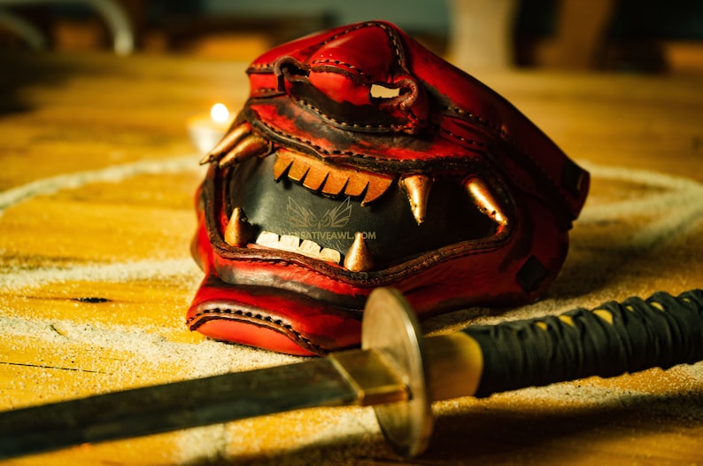 K&ouml;nnte beinhalten: Eine rote Leder-Samurai-Maske mit goldenen Akzenten und scharfen Z&auml;hnen, die auf einer Holzoberfl&auml;che neben einem schwarzen und goldenen Katana-Schwert liegt.