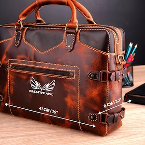 May include: A brown leather briefcase with a front pocket and a buckle closure. The briefcase has a logo that says "CREATIVE AWL" and an owl design. The briefcase measures 16 inches wide, 11 inches tall, and 3.1 inches deep.