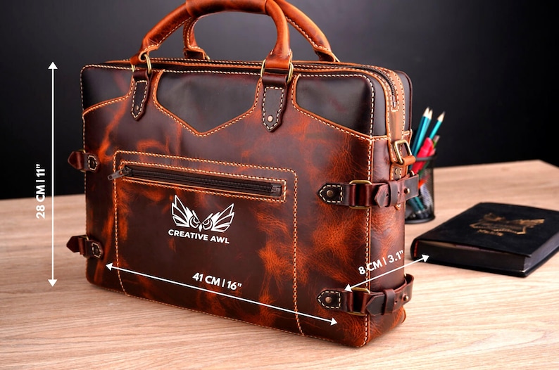 May include: A brown leather briefcase with a front pocket and a buckle closure. The briefcase has a logo that says "CREATIVE AWL" and an owl design. The briefcase measures 16 inches wide, 11 inches tall, and 3.1 inches deep.