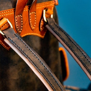 May include: Close-up of a brown leather backpack with two adjustable straps. The straps are stitched with brown thread and have a brass buckle.