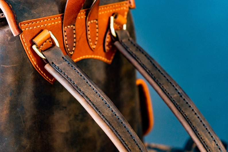May include: Close-up of a brown leather backpack with two adjustable straps. The straps are stitched with brown thread and have a brass buckle.
