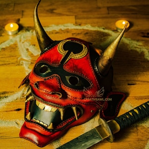 K&ouml;nnte beinhalten: Eine rote und schwarze Leder-Oni-Maske mit goldenen Akzenten und H&ouml;rnern. Die Maske liegt auf einer Holzoberfl&auml;che mit einem Sandkreis darum herum. Ein Katana-Schwert mit einem schwarzen und goldenen Griff liegt auf der Oberfl&auml;che.