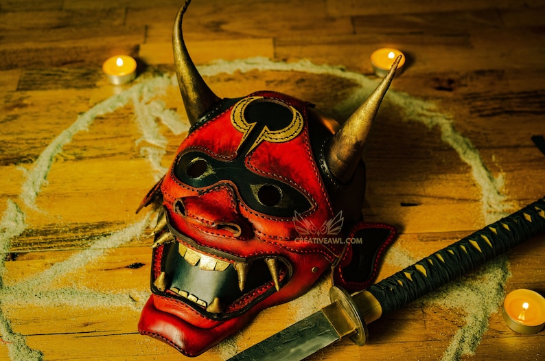 K&ouml;nnte beinhalten: Eine rote und schwarze Leder-Oni-Maske mit goldenen Akzenten und H&ouml;rnern. Die Maske liegt auf einer Holzoberfl&auml;che mit einem Sandkreis darum herum. Ein Katana-Schwert mit einem schwarzen und goldenen Griff liegt auf der Oberfl&auml;che.