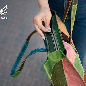 May include: A multi-coloured patchwork tote bag featuring panels of green, pink, and brown suede leather with visible stitching. The bag includes a top zip closure and two leather shoulder straps. The logo CREATIVE AWL is visible in the corner.
