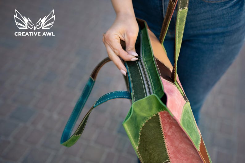 May include: A multi-coloured patchwork tote bag featuring panels of green, pink, and brown suede leather with visible stitching. The bag includes a top zip closure and two leather shoulder straps. The logo CREATIVE AWL is visible in the corner.