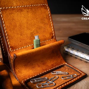 May include: A handcrafted, brown leather phone stand with a built-in charging port and a small tray for paperclips. The stand has a stitched design and a green charging cable. A smartphone and notebook are visible in the background.