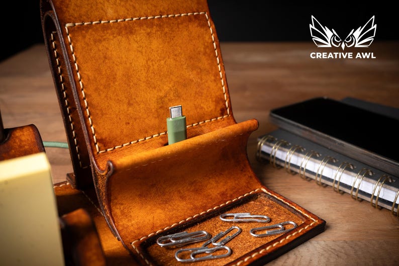 May include: A handcrafted, brown leather phone stand with a built-in charging port and a small tray for paperclips. The stand has a stitched design and a green charging cable. A smartphone and notebook are visible in the background.
