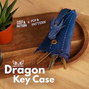 May include: Blue leather dragon key case with stitching detail. The case is open and has keys inside. The text "Dragon Key Case" is visible below the case. The text "DXF & PDF PATTERN" is visible above the case.