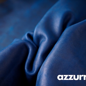 May include: Close-up of a deep blue leather fabric with a textured surface. The fabric is folded and draped, showcasing its soft and supple texture. The word "azzurro" is printed in white at the bottom of the image.