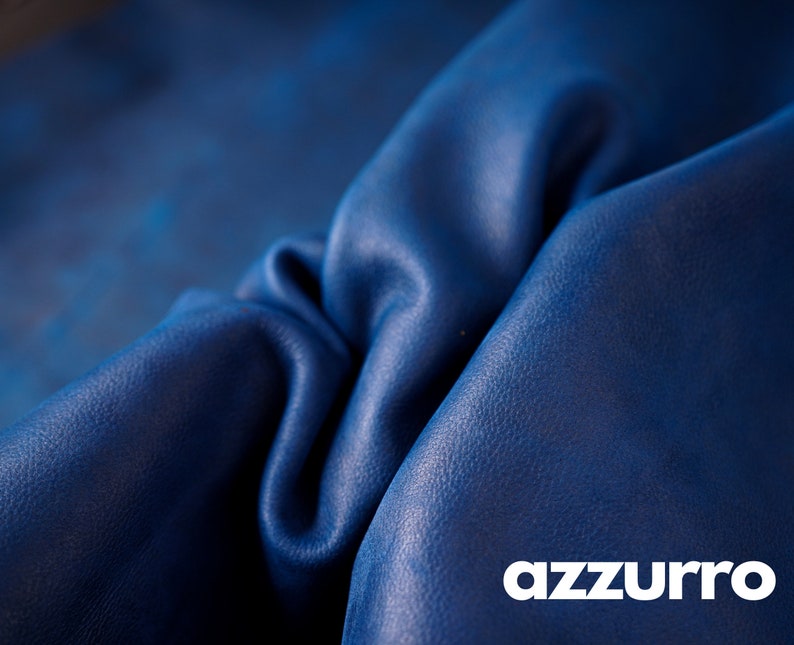 May include: Close-up of a deep blue leather fabric with a textured surface. The fabric is folded and draped, showcasing its soft and supple texture. The word "azzurro" is printed in white at the bottom of the image.