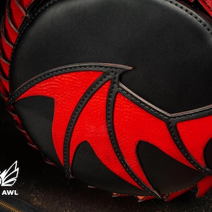 May include: A black and red round handbag with a bat wing design. The bag has a black body with red accents and stitching. A gold-coloured ring is visible on the side. The Creative Awl logo is at the bottom.