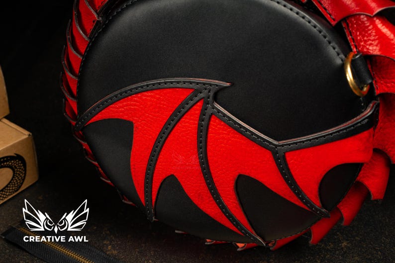 May include: A black and red round handbag with a bat wing design. The bag has a black body with red accents and stitching. A gold-coloured ring is visible on the side. The Creative Awl logo is at the bottom.