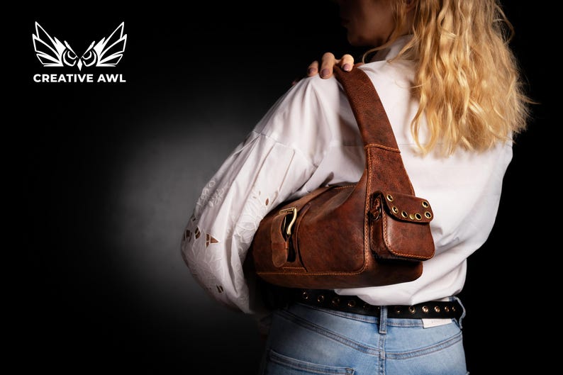 Puede incluir: Un bolso de hombro de cuero marr&oacute;n con cierre de solapa y un peque&ntilde;o bolsillo frontal. El bolso tiene herrajes dorados y lo lleva una persona con el pelo largo y rubio.