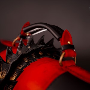 Op de afbeelding: Een rode en zwarte leren tas met een zwarte handvat en gouden accenten. De tas heeft een draakachtig ontwerp met spikes.