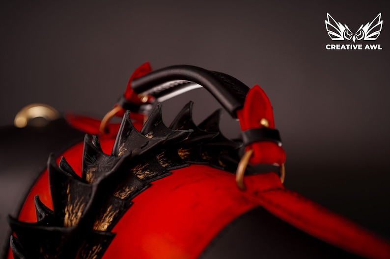 Op de afbeelding: Een rode en zwarte leren tas met een zwarte handvat en gouden accenten. De tas heeft een draakachtig ontwerp met spikes.