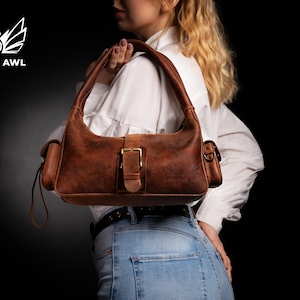 Puede incluir: Un bolso de mano de cuero marr&oacute;n con una hebilla dorada y una correa. El bolso tiene dos bolsillos a los lados y un peque&ntilde;o bolsillo en la parte delantera.