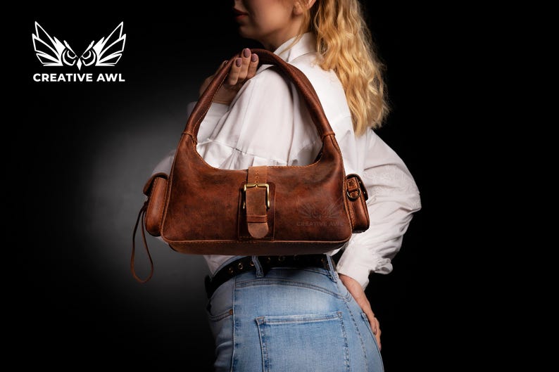 Puede incluir: Un bolso de mano de cuero marr&oacute;n con una hebilla dorada y una correa. El bolso tiene dos bolsillos a los lados y un peque&ntilde;o bolsillo en la parte delantera.
