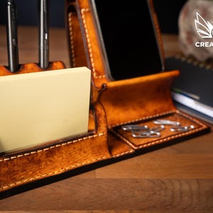 May include: A handcrafted, brown leather desk organizer with compartments for a phone, pens, sticky notes, and paper clips. The organizer has a stitched design and a warm, natural finish. The Creative Awl logo is visible in the background.