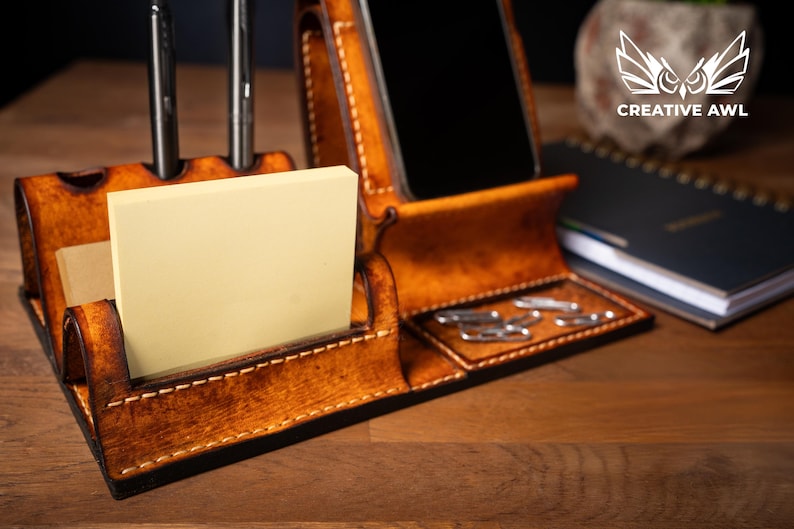 May include: A handcrafted, brown leather desk organizer with compartments for a phone, pens, sticky notes, and paper clips. The organizer has a stitched design and a warm, natural finish. The Creative Awl logo is visible in the background.