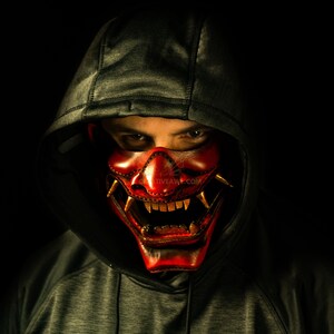 K&ouml;nnte beinhalten: Eine Person tr&auml;gt einen dunkelgrauen Kapuzenpullover mit einer roten und goldenen Oni-Maske. Die Maske hat scharfe Z&auml;hne und H&ouml;rner.