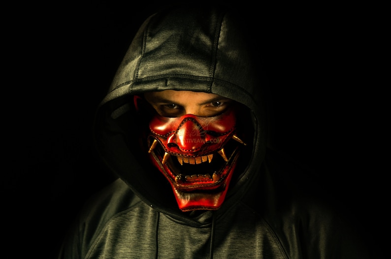 K&ouml;nnte beinhalten: Eine Person tr&auml;gt einen dunkelgrauen Kapuzenpullover mit einer roten und goldenen Oni-Maske. Die Maske hat scharfe Z&auml;hne und H&ouml;rner.
