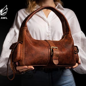 Puede incluir: Un bolso hobo de cuero marr&oacute;n con cierre de hebilla y un peque&ntilde;o bolsillo lateral. El bolso tiene una correa larga que se puede llevar al hombro.
