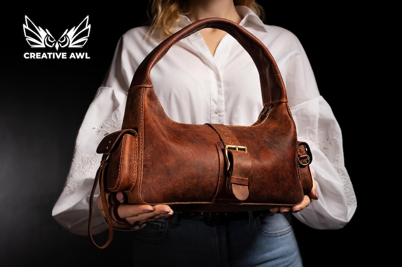 Puede incluir: Un bolso hobo de cuero marr&oacute;n con cierre de hebilla y un peque&ntilde;o bolsillo lateral. El bolso tiene una correa larga que se puede llevar al hombro.