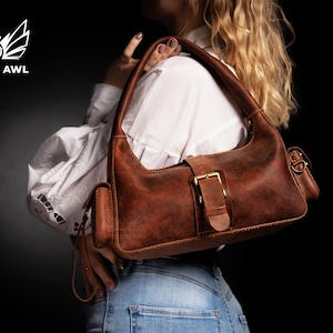 Puede incluir: Un bolso de cuero marr&oacute;n con cierre de hebilla y un peque&ntilde;o bolsillo lateral. El bolso se lleva sobre el hombro de una persona que lleva una camisa blanca y pantalones vaqueros azules.
