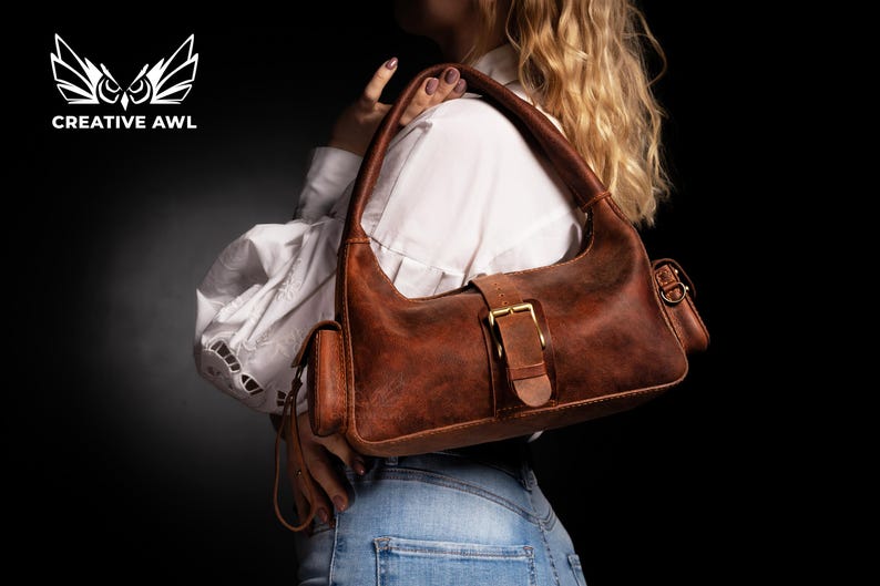 Puede incluir: Un bolso de cuero marr&oacute;n con cierre de hebilla y un peque&ntilde;o bolsillo lateral. El bolso se lleva sobre el hombro de una persona que lleva una camisa blanca y pantalones vaqueros azules.