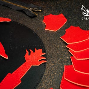 May include: A leather craft project is shown, featuring a black leather base with red leather cutouts. These include a stylized hand and a star-like shape. Red shield-shaped pieces are scattered around. A zipper is also visible in the image.