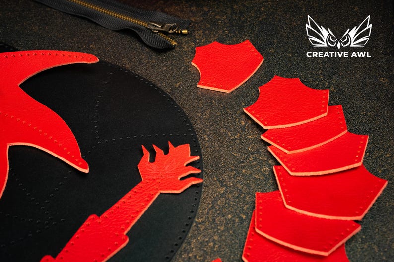May include: A leather craft project is shown, featuring a black leather base with red leather cutouts. These include a stylized hand and a star-like shape. Red shield-shaped pieces are scattered around. A zipper is also visible in the image.