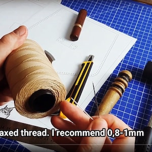 Może przedstawiać: Zbliżenie na szpulkę woskowanej nici, narzędzie do obr&oacute;bki sk&oacute;ry i młotek na niebieskiej macie do cięcia. Tekst "and a waxed thread. I recommend 0,8-1mm" jest widoczny na zdjęciu.