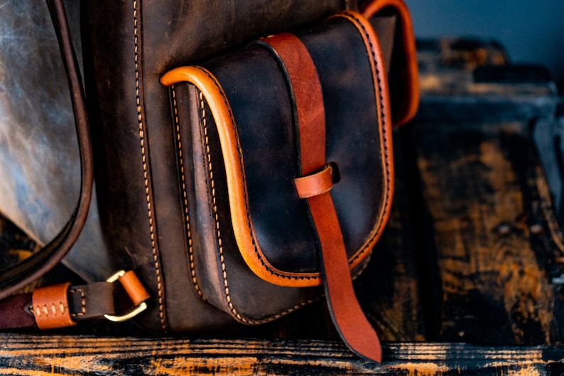 May include: Close-up of a brown leather backpack with a dark brown leather pocket and orange stitching. The pocket has a brown leather strap with a buckle.