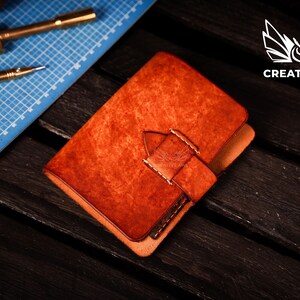May include: A brown leather notebook with a flap closure and a stitched strap. The notebook is on a dark wood surface.