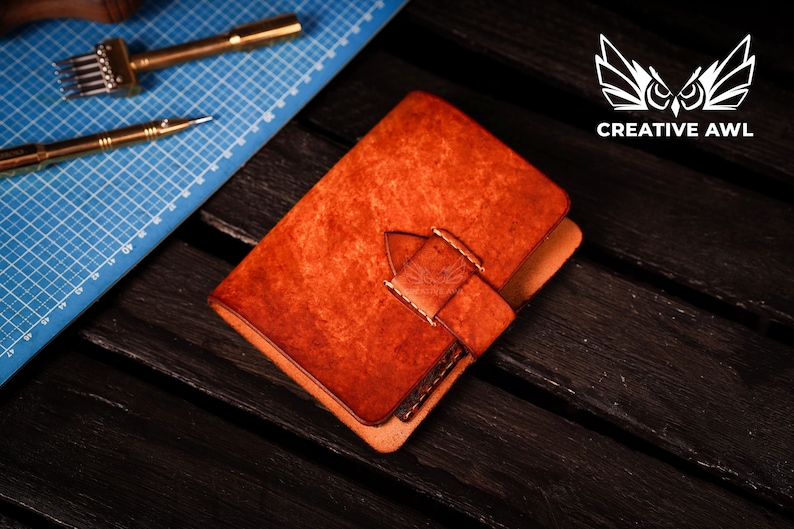 May include: A brown leather notebook with a flap closure and a stitched strap. The notebook is on a dark wood surface.