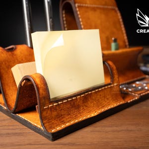 May include: A handcrafted leather desk organizer in shades of brown, featuring compartments for a phone, sticky notes, and pens. The organizer has a curved design and visible stitching, with the "CREATIVE AWL" logo in white.