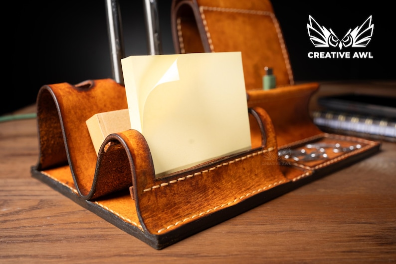 May include: A handcrafted leather desk organizer in shades of brown, featuring compartments for a phone, sticky notes, and pens. The organizer has a curved design and visible stitching, with the "CREATIVE AWL" logo in white.