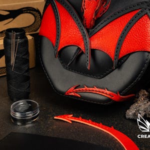 Op de afbeelding: Een zwart en rood lederen tas met een drakenvleugelontwerp, vergezeld door een klos zwart garen, naainaalden, een klein potje zwarte was en losse rode lederen uitsparingen, met het Creative Awl logo zichtbaar in de hoek.