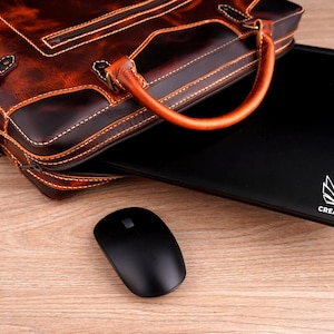 May include: A brown leather briefcase with a laptop computer inside. The laptop has a white owl logo with the text "CREATIVE AWl" on the back. A black wireless mouse is on a wooden surface.